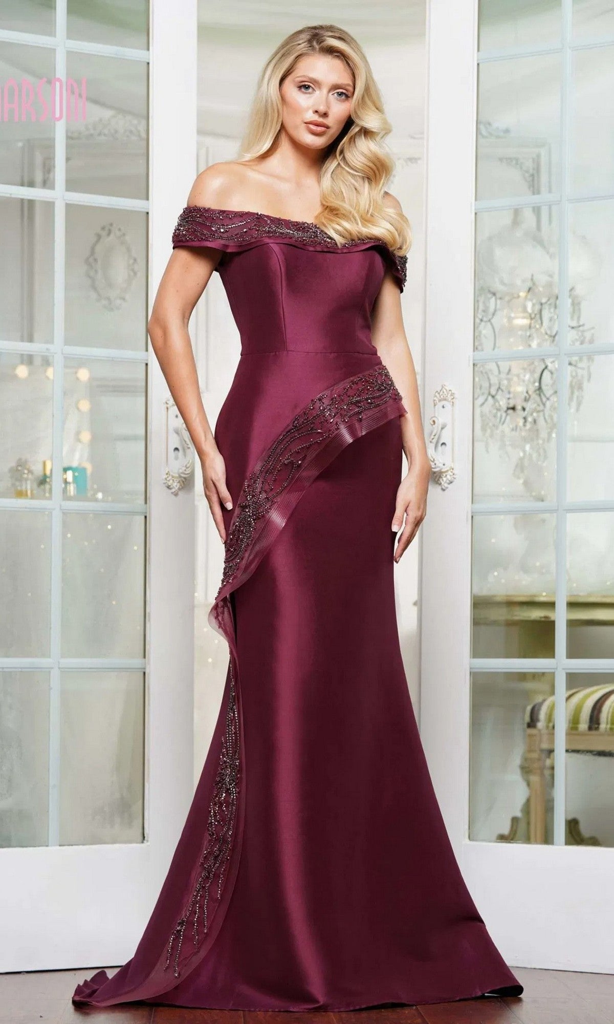 A woman stands indoors wearing the Marsoni MV1332 Long Formal Dress in wine, featuring an off-shoulder design, beaded accents, a slight train, and a refined mermaid silhouette.